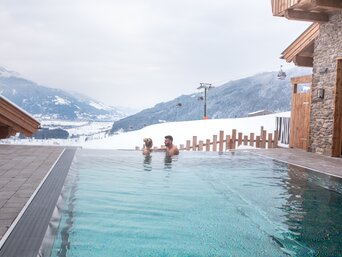 Paar entspannt im beheizten Außenpool des nachhaltigen Hotel-Dorfs Zaglgut mit Panoramablick auf die verschneite Berglandschaft – mit Heiz- und Lüftungstechnik sowie Gebäudeautomation von bösch. | © Hotel & Chalets Zaglgut