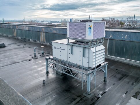 Mit nachhaltigen Technologien etablierte das Erfolgsduo INFRANORM® & bösch neuartige Lösungen. Hallenluft-Absaugung, Frischluftzufuhr und Luftverteilung sind intelligent kombiniert. | © INFRANORM® Luftaufnahme eines großen, auf einem Fabrikdach installierten Lüftungssystems. Die Anlage steht auf einer Metallstruktur und ist vorwiegend in Weiß gehalten, mit einem oberen Gitter in Silber. Das Dach ist nass, möglicherweise nach einem Regenschauer. Im Hintergrund ist eine städtische Umgebung mit entfernten Gebäuden und leicht bewölktem Himmel zu erkennen. | © INFRANORM®