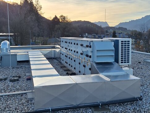Das auf dem Dach installierte RLT-Gerät von bösch versorgt den Kindergarten mit perfekt konditionierter Raumluft. Ein Dach mit mehreren Klimaanlagen-Einheiten in verschiedenen Größen und Formen bei Sonnenuntergang. Die Anlagen sind auf einem mit Kies bedeckten Dach platziert, das sich vor einem Hintergrund mit bewaldeten Bergen erstreckt. Der Himmel ist leicht bewölkt und färbt sich orange durch die untergehende Sonne.